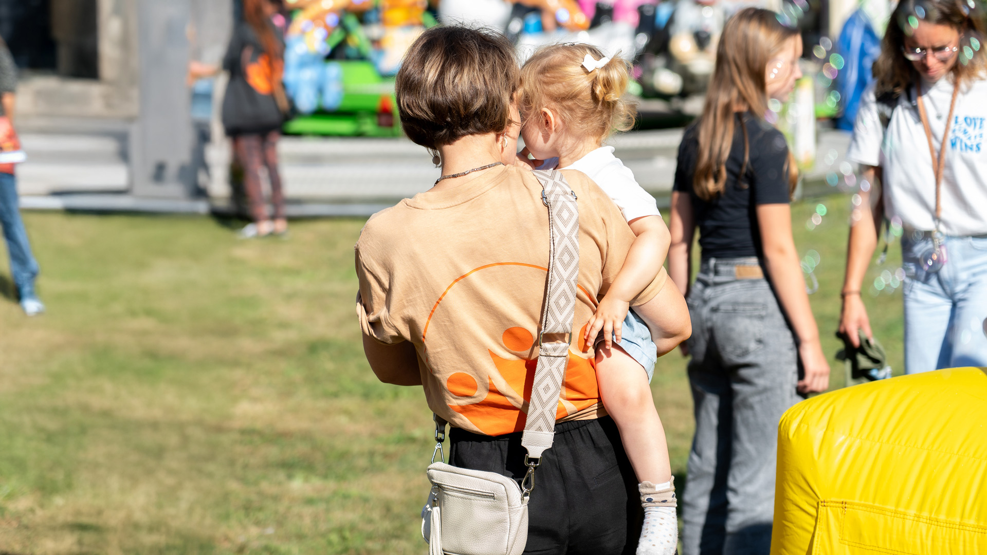 Foto einer Mitarbeiterin, wie sie ein Kind bei einer Veranstaltung hält. Im Hintergrund sind mehr Personen und es fliegen Seifenblasen.